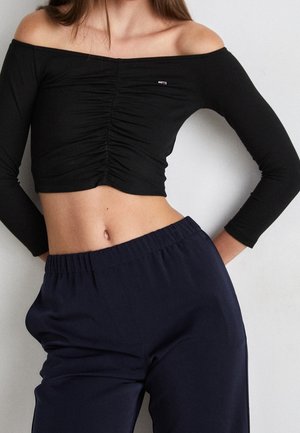 Kobieta ubrana w czarny marszczony, długi crop top z odsłoniętymi ramionami oraz luźne granatowe spodnie stojąca na tle gładkiej białej ściany.