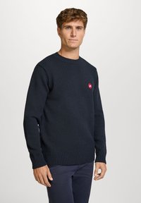 Marineblå strikket genser med rund hals, ribbestrikkede mansjetter og nederkant, med en liten rød logo på brystet. Myk tekstur.
