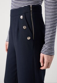 Pantalons taille haute marine avec plis et accents de boutons en argent. Présente une fermeture éclair sur le côté, fabriqué à partir d'un tissu texturé.