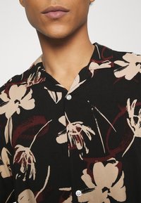 Lyhythihainen paita, jossa musta pohja ja suuret beiget sekä viininpunaiset kukkakuviot, lovikaulus ja etupuolella nappikiinnitys.