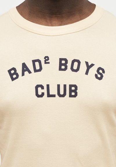 Κοντινό πλάνο μιας μπεζ ριμπ μπλούζας με την επιγραφή «BAD² BOYS CLUB» τυπωμένη με έντονα μαύρα γράμματα στο στήθος.