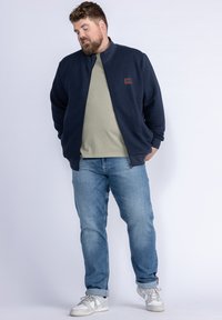 Navy textured zip-up jacket with stand collar over a light green T-shirt, paired with blue jeans and white sneakers.