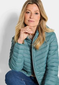 Lichtblauwe pufferjack met horizontale en diagonale quiltpatronen, voorrits en hoge kraag. Draag het over een beige trui.