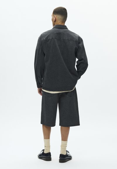 Tamno plava traper jakna s dugim rukavima i džepovima na prsima uparena s traper šortsem do koljena. Završeno svijetlim čarapama i crnim cipelama.