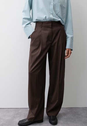 Persona che indossa pantaloni marroni larghi, scarpe nere e una camicia azzurro chiaro a manica lunga abbottonata, con una mano in tasca.