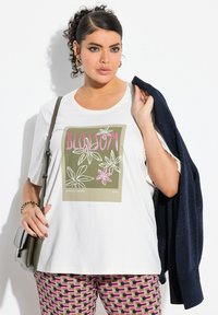 Camiseta blanca de manga corta con un gráfico floral en verdes, rosas y blancos, combinada con pantalones estampados en rosa y morado y un cárdigan oscuro.