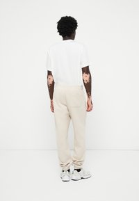 Beige mjukisbyxor med sidficka och elastisk midja; matchade med en vit t-shirt och grå träningsskor, sedd bakifrån.