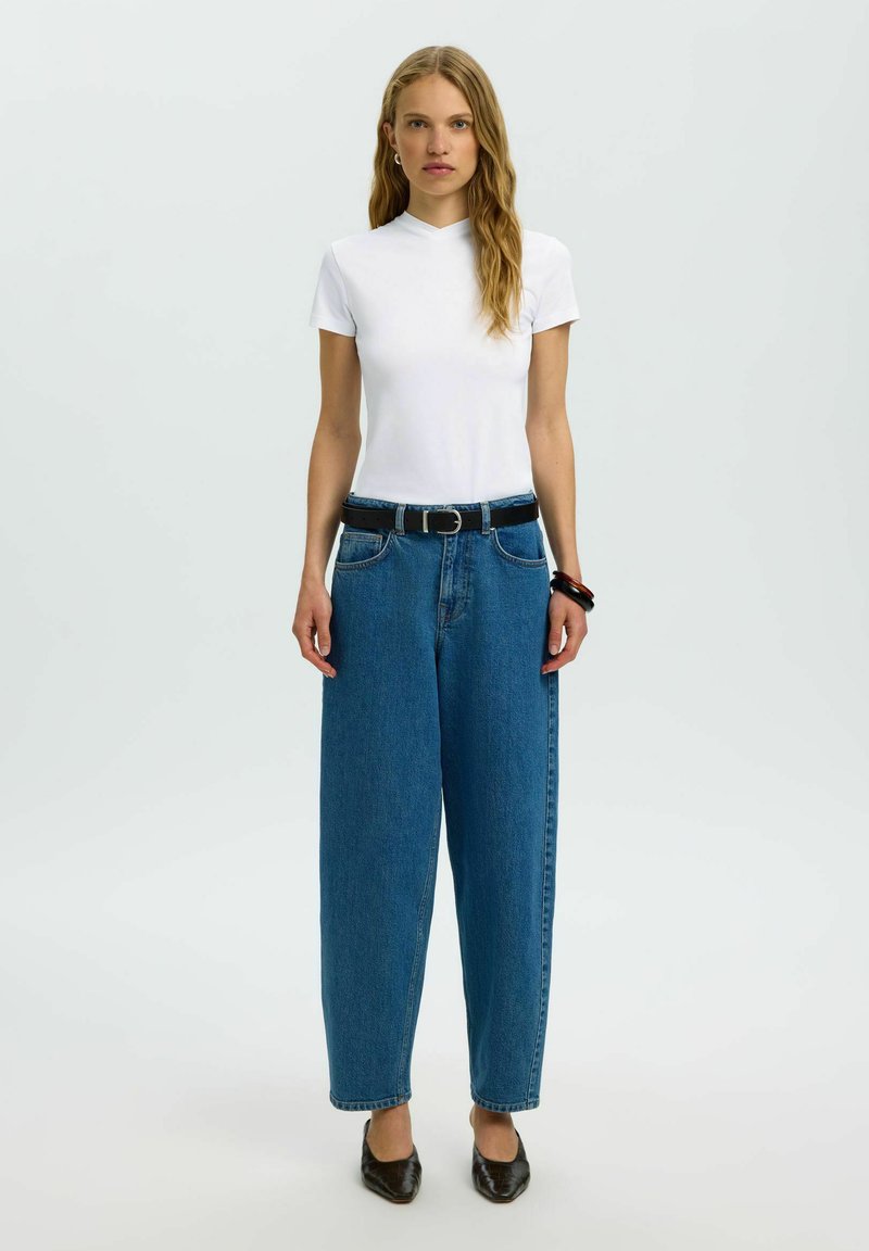 Jeune femme debout portant un haut blanc à manches courtes, un jean large bleu avec une ceinture noire, des chaussures noires et des bracelets au poignet gauche.