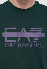 Tumši zaļš kokvilnas T-krekls ar purpursarkanu grafiku, kas satur tekstu "EA" un "EMPORIO ARMANI". Klasiska apaļa kakla izgriezums, īsas piedurknes.