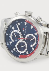 Sølv rustfritt stål kronograf klokke med en mørk blå urskive, røde detaljer, tre subskiver og hvite timemarkører. Har en metallarmbånd.