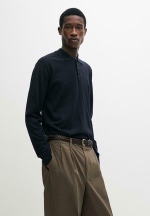 Uomo che indossa una polo nera a maniche lunghe infilata in pantaloni marroni a vita alta con pieghe e cintura intrecciata, mani nelle tasche, sfondo neutro.