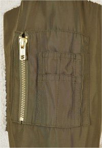 Tissu vert olive avec une fermeture éclair dorée sur le côté et une poche à patch. Le matériau a une texture lisse avec de subtiles lignes diagonales.