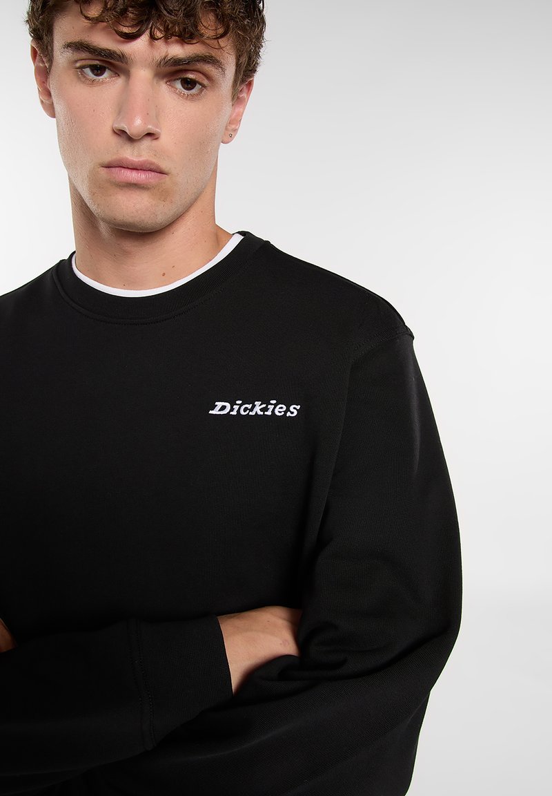 Zwarte trui met geribde halslijn en manchetten, met wit geborduurd "Dickies"-logo op de linkerborst. Zachte katoenmengstof.