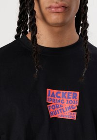 Melns kokvilnas T-krekls ar daudzkrāsainu tekstu sarkanā un zilā krāsā, uz kura ir uzraksts "JACKER SPRING 2025 FOREVER HUSTLING" priekšā.