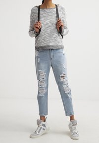 Grau-weißer melierter Pullover mit weitem Ausschnitt, verwaschene hellblaue Jeans mit großen Rissen und weiße High-Top-Sneaker. Schwarze Rucksackriemen.