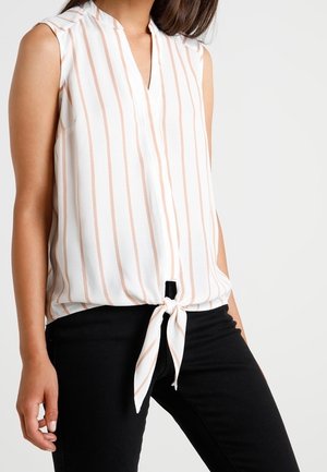 Mouwloze witte blouse met verticale beige strepen, V-hals en een knoop aan de voorkant, gedragen met een zwarte broek door een staande persoon.