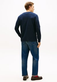 Camicia a maniche lunghe blu navy con scollo rotondo, abbinata a jeans denim azzurri. La modella è di spalle, mostrando il design semplice e la vestibilità.