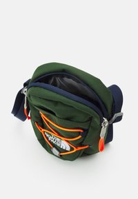 Zielona torba typu crossbody z pomarańczowymi akcentami, zamkiem błyskawicznym, przednim sznurkiem bungee i logo. Zawiera regulowany granatowy pasek.