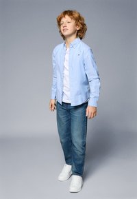 Camisa de botão azul clara sobre uma t-shirt branca, combinada com jeans azuis e ténis brancos. O conjunto apresenta um pequeno detalhe de logótipo na camisa.