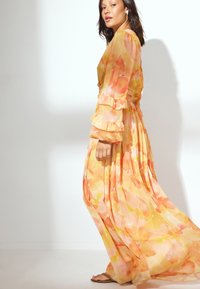 Frau in einem fließenden gelb- und orangefarbenen Blumenkleid, die barfuß an einer weißen Wand mit einem sanften Schatten steht.