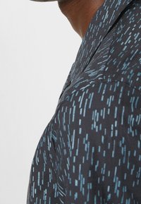 Zbliżenie na ramię i szyję osoby noszącej ciemną koszulę w niebieskie, pionowe paski na jasnym tle.