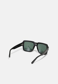 Ochelari de soare pătrați negri cu lentile verde închis, ramă groasă din plastic și brațe subțiri. Prezintă un accent metalic lângă balama.