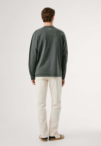 Camisa gris de manga larga con corte relajado y cuello redondo, combinada con pantalones de color crema y zapatos claros. Sin patrones ni detalles notables visibles.