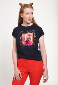 Mörkblå t-shirt med en färgglad Miss Piggy-grafik. Designen inkluderar texten: "MISS PIGGY FABULOUS" i rosa. Modellen bär röda byxor.