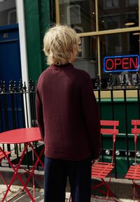 Pull en ribbed bordeaux avec un col montant, vu de dos. Présente une coupe décontractée, associé à un pantalon bleu marine et une table de bistrot en métal rouge à proximité.