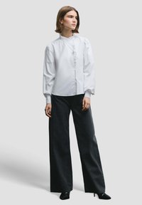 White long-sleeve shirt with a high collar and ruffled front, paired with wide-leg dark pants featuring small metallic accents.