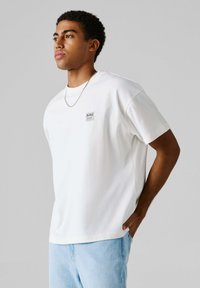 Camiseta blanca de algodón con un corte holgado, que presenta un pequeño parche de logo en blanco y negro en el pecho y mangas cortas.