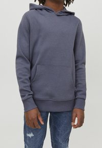 Sudadera con capucha en gris oscuro, fabricada en tejido texturizado. Presenta un bolsillo canguro frontal y puños acanalados. Se lleva con unos vaqueros desgastados de color azul.