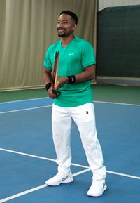 Zelena majica kratkih rukava za tenis, bijele hlače i bijele tenisice s crnim detaljima. Drži reket za tenis na unutarnjem terenu.