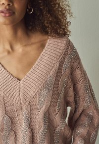 Rosa gestrickter Pullover mit V-Ausschnitt, der über Zopfmuster und silberne Verzierungen an den Seiten verfügt. Strukturierter Stoff.