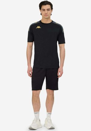 T-shirt de sport noire à manches courtes, arborant des accents de logo dorés sur les épaules, associée à un short noir et des baskets de couleur claire.