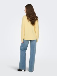 Pull jaune oversize, jean bleu ample, chaussures noires à talons, longs cheveux bruns ondulés, vue de dos.