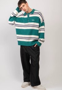 Zeleno-beli črtasti oversized pleteni pulover s ovratnikom, opremljen z majhnim logotipom. Kombiniran z ohlapnimi črnimi hlačami in belimi supergami.