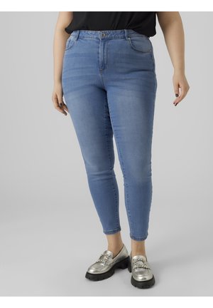 Personne portant un jean skinny bleu clair taille moyenne et des mocassins argentés métalliques avec des boucles décoratives, debout sur un fond uni.
