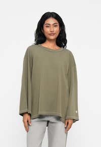 Χακί πουκάμισο με μακριά μανίκια και χαλαρή εφαρμογή, στρογγυλή λαιμόκοψη, κατεβασμένους ώμους και ίσιο στρίφωμα. Απλή ραφή και τονισμένο λογότυπο.