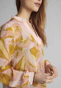 Lätt, transparent blus i rosa med gula och gyllene botaniska mönster. Har en notchad krage och volangärmar med knappaccenter.