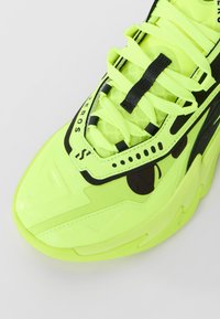 Nærbillede af en neongul sneakersnude med sorte detaljer, tykke snørebånd og markante syninger på en lys baggrund.