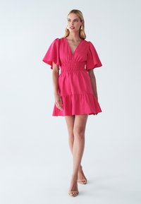 Rosa kurzes Kleid mit V-Ausschnitt, Puffärmeln und einem gesmokten Taillenbund. Es hat einen ausgestellten Rock und eine strukturierte Stoffoberfläche.
