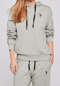 Grå hoodie i mjukt material, med en framficka, svarta dragskor och en liten svart logotyp på bröstet.