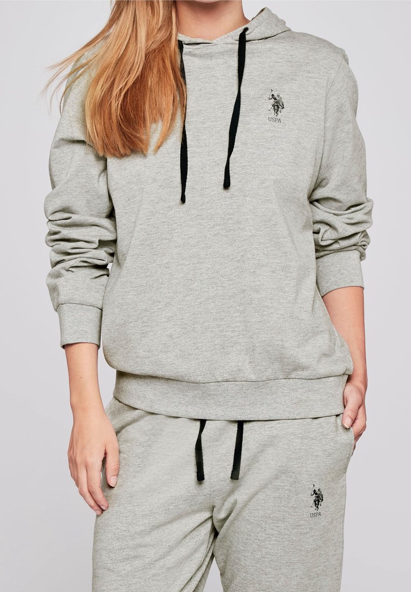 Grå hoodie i mjukt material, med en framficka, svarta dragskor och en liten svart logotyp på bröstet.