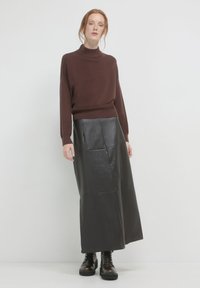 Frau in einem braunen Rollkragenpullover, langer schwarzer Lederröhre und schwarzen Schnürstiefeln, die vor einem schlichten weißen Hintergrund steht.