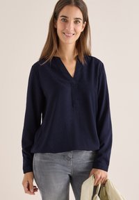 Mørkeblå bluse med lange ærmer, V-hals og subtil tekstur. Båret sammen med grå jeans og holder en lys håndtaske.