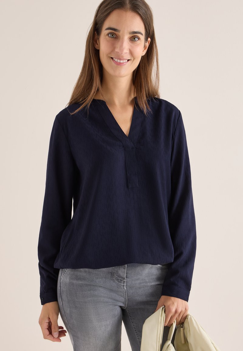 Mørkeblå bluse med lange ærmer, V-hals og subtil tekstur. Båret sammen med grå jeans og holder en lys håndtaske.