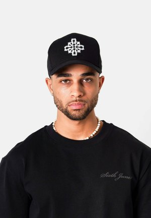 Man met getrimde baard draagt zwarte pet met wit kruisdesign, zwart shirt en parelketting tegen een grijze achtergrond.