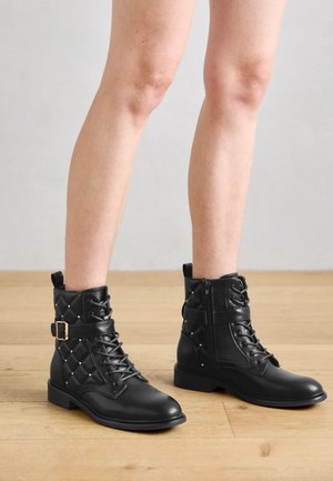 Ben iført sorte snørestøvler til ankler med quiltede sider, små gyldne nitter, sidelås og rem med spænde på træbundsbaggrund.