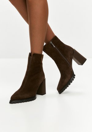 Stivali ankle in suede marrone con punta a punta, tacco a blocco e cerniere laterali; suole in gomma texture offrono aderenza.